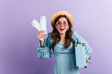 Close-up portrait of contented girl in sunglasses on lilac background. Woman shows tickets for upcoming trip
