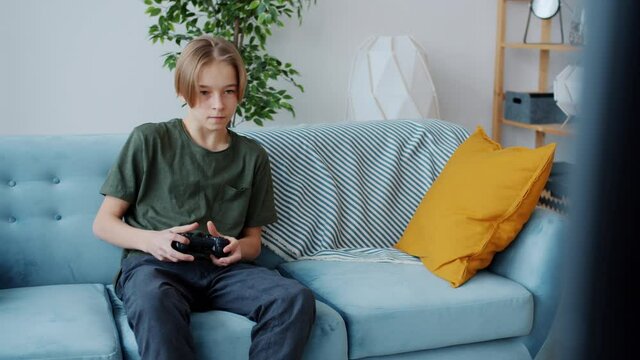 Cheerful teenage boy is playing video game using controller pushing buttons and smiling sitting on sofa at home. Contemporary devices and adolescence concept.