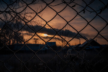 a bright disc of the sun peeps through the clouds against the background of a lattice of metal mesh on the fence