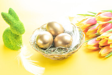 Basket easter decoration: Golden eggs in basket with spring tulips, white feathers on pastel yellow background. Congratulatory easter design.