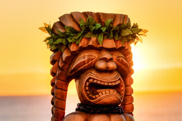 Hawaiian Tiki Statue with an amazing sunrise in the background. The tiki is dressed up with traditional leaves and a macadamia necklace. It is made of wood and looks large in size. - obrazy, fototapety, plakaty