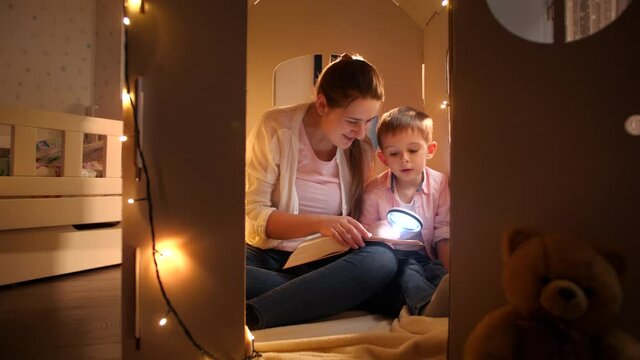 Happy smiling boy with young mother sitting in tent or toy cardboard house and reading book with flashlight at night. Concept of child education and family having time together at night