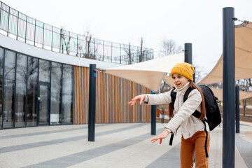 Preteen girl dressed yellow hat, jacket, backpack and trousers walking outdoor in the street of the city