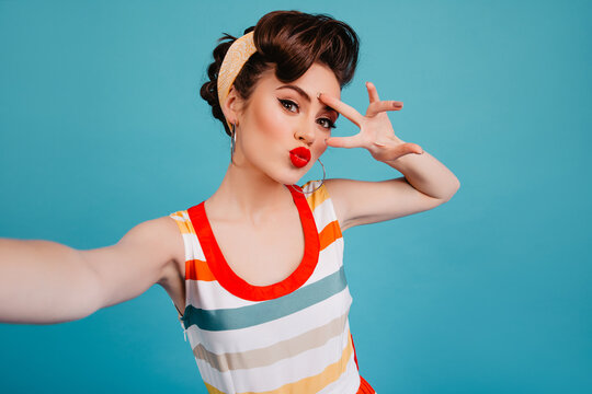 Gorgeous pinup girl having fun on blue background. Good-looking woman in striped dress taking selfie with peace sign.
