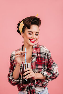 Studio shot of pinup lady isolated on pink background. Laughing girl in checkered shirt drinking soda.