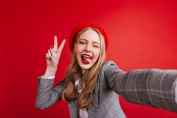 Carefree girl in elegant jacket taking selfie. Funny french woman showing peace sign on red background.