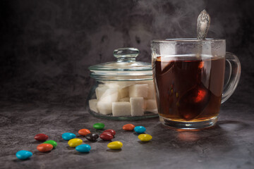 Tea in a glass cup with a spoon, and a glass sugar bowl with a lid and lump sugar and candy on a gray background