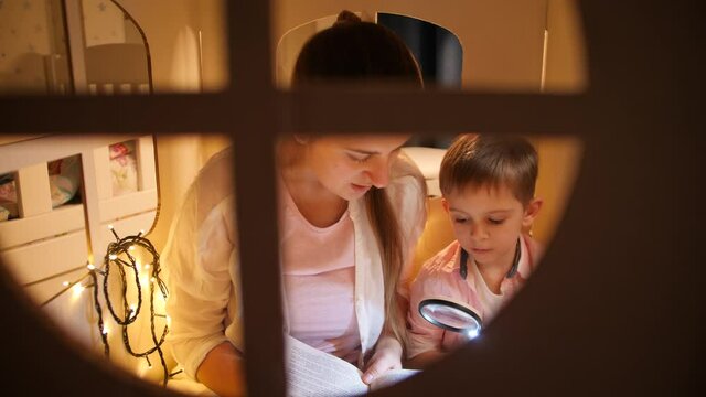 Portrait Of Cute Boy With Young Mohter Reading Big Book In Tent Or Toy House Before Going To Sleep. Concept Of Child Education And Family Having Time Together At Night