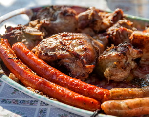 Charcoal-fried chicken pieces and sausages laid out on a plate on a background of oilcloth