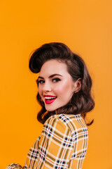 Charming woman with curly hairstyle looking over shoulder. Smiling pinup girl in checkered shirt posing on yellow background.