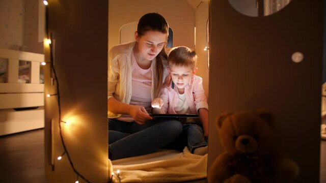 Little smiling boy with young mother playing in tent or toy house and using tablet computer. Concept of child education and family having time together at night