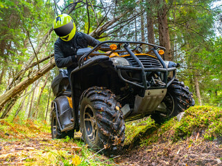 Portrait of a biker on an ATV. Biker rides in woods. He looks at wheels of quadbike. Biker on the background of the forest. ATV rider in green taiga. ATV as a symbol of extreme sports.