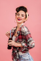 Appealing pinup woman in checkered shirt smiling at camera. Studio shot of attractive brunette girl isolated on pink background with bottle.