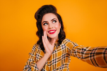 Glamorous young woman in checkered shirt taking selfie. Studio shot of amazing pinup girl isolated on yellow background.