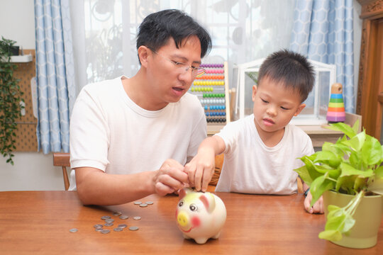 Father and son putting Thai coin into piggy bank at home, Happy family money savings concept. Dad teaching son on savings & financial planning, Little 5 years old kid saving money for future concept