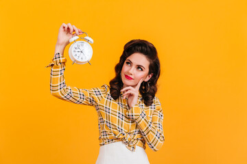 Beautiful pensive woman holding clock. Studio shot of playful pinup girl posing on yellow background.
