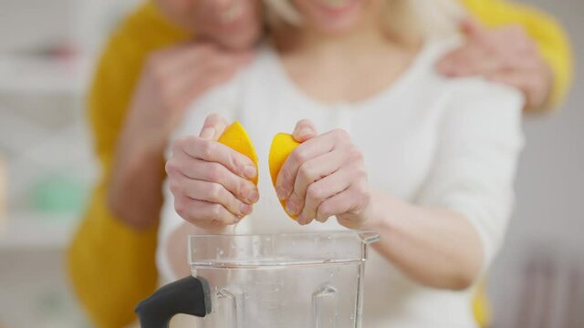 Close-up of female hands squeezing lemon in blender with blurred unrecognizable couple hugging at background. Cheerful man showing strength gesture as smiling woman preparing vegan healthy drink.