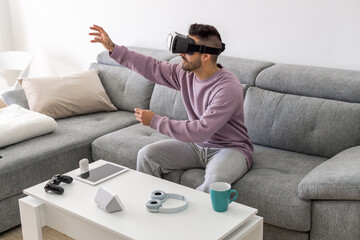 A handsome boy plays a game inside virtual reality wearing virtual reality glasses while on the couch in his living room.