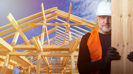Fototapeta premium The Builder holds up the boards and smiles at the camera. A man in a construction helmet on the background of a wooden frame of a cottage. High-quality lumber. Construction of country houses.