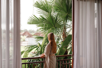 Beautiful young woman stand  on balcony terrace of jungle villa with morning coffee, wearing...