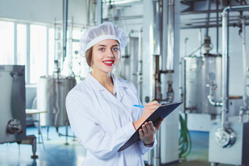 A young beautiful girl in white overalls makes notes in a tablet on the background of equipment of a food processing plant. Quality control in production