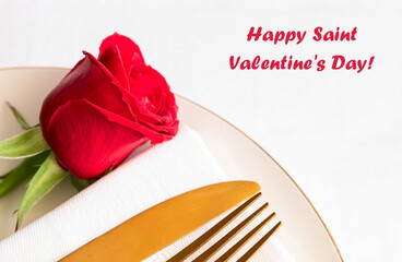 Festive table setting in white tone. Plate, gold cutlery, napkin and red rose on white tableware in restaurant interior. Saint valentines day celebration or romantic dinner concept.