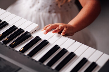 close-up, the hand of a five-year-old girl in a white dress presses her fingers on the keys of an electronic synthesizer in the living room.