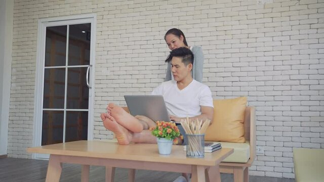 Young asian man sitting on the couch work with laptop while woman massaging to him in the luxury contemporary living room next the window at home. Filling their home with love and laughter.