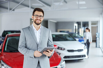 Smiling friendly car seller in suit standing in car salon and holding tablet. It's always pleasure...