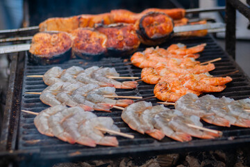 Cooking tiger shrimp, prawn and salmon skewers on grill at summer local food market - close up. Outdoor cooking, barbecue, gastronomy, seafood, cookery, street food concept