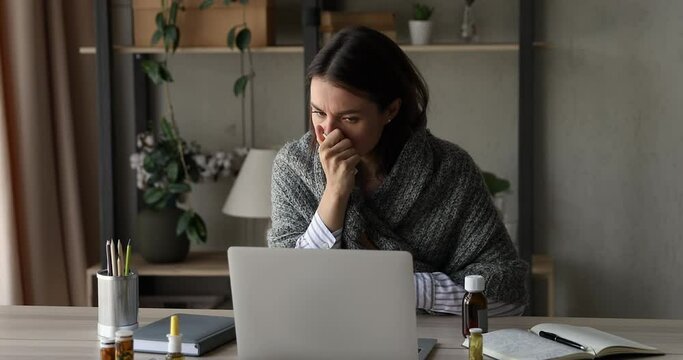 Unhealthy young businesswoman using paper tissue, sitting at workplace covered in warm plaid, working on computer at home office suffering from first flu grippe symptoms, self-isolation during illness