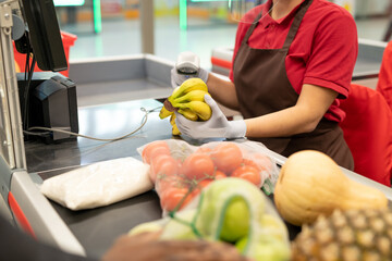 Young female cashier in uniform and gloves holding bunch of bananas