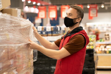 Young male worker of supermarket unpacking huge stack of packed boxes with goods