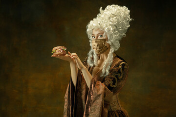 Portrait of medieval young woman in vintage clothes, golden face mask going to eat burger on dark background. Royal person protected from covid. Concept of comparison of eras, modern, fashion.