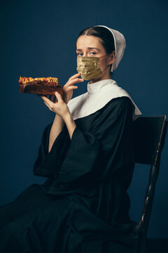 Medieval Young Woman As A Nun In Vintage Clothing And Golden Face Mask With Fast Food On Dark Blue Background. Concept Of Comparison Of Eras, Fashion, Ad, Healthcare. Royal Person Protected From Covid