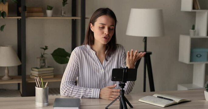 Concentrated millennial beautiful female blogger sitting at table in front of telephone on tripod stabilizer, recording educational video on mobile web camera at home office, sharing knowledge.