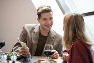 Guy with smartphone looking at his girlfriend with smile while talking to her