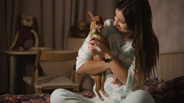 Young girl is sitting on the bed in the bedroom with small dog. Small pet and its owner 