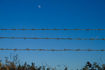 Three lines of barbed wire running horizontally across the photo against blue sky with moon in the background.