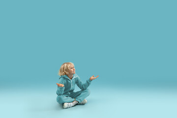 Sitting, wondered. Childhood and dream about big and famous future. Pretty curly boy isolated on blue studio background. Childhood, dreams, imagination, education, facial expression, emotions concept.