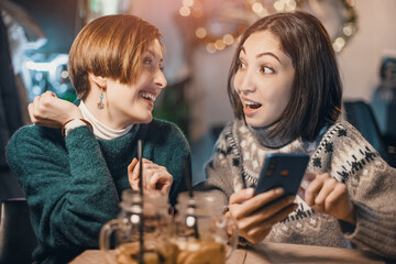 Two happy women sit in a cafe, chat with each other and watch the news on social networks on their mobile phone. The concept of relationships and communication
