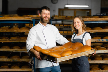 Bakers at the bakery. A man and a woman are holding a tray with fresh hot bread in the background of shelves with bread in a bakery. Industrial bread production