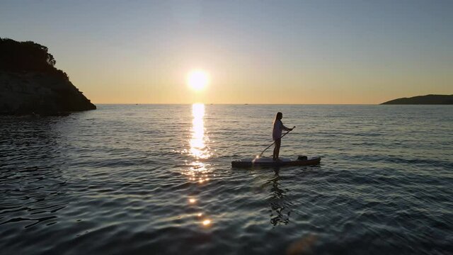 Silhouette of woman rowing SUP-board with paddle along sea with rippling waves against clear sky at sunset reflecting on water
