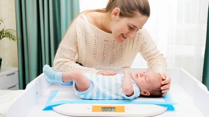 Smiling mother looking and stroking her little son while weighing him on digital scales on changing table at home. Concept of babies and newborn hygiene and healthcare. Caring parents with little