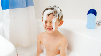 Cheerful little boy playing in bath and splashing water while washing his head with shampoo....