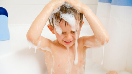 Cute little boy sitting in bath and washing hair with shampoo