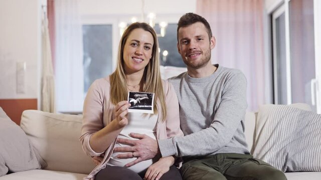 Happy young pregnant couple sits at home on couch and showing ultrasonic picture towards the cam shot in 4k