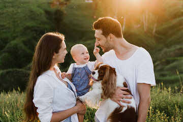 Young beautiful family with a little daughter and a dog hug, kiss and walk in nature at sunset.