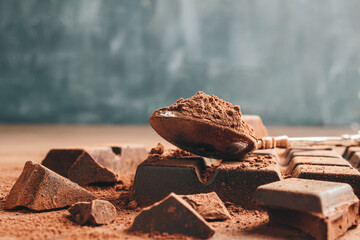 Brown cocoa powder in the spoon and chopped chocolate cubes, on dark background with copy space