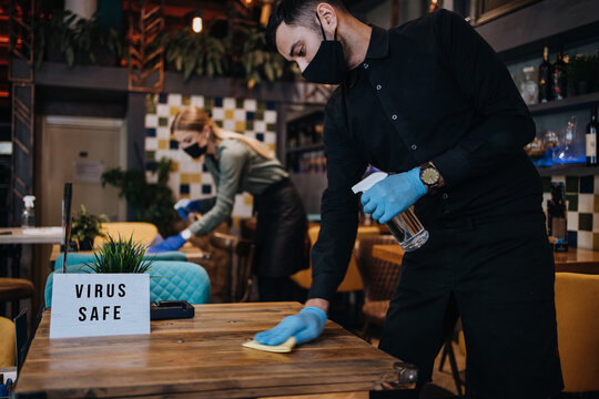 Young restaurant workers waiters cleaning and disinfecting tables and surfaces against Coronavirus pandemic disease. They are wearing protective face masks and gloves.
